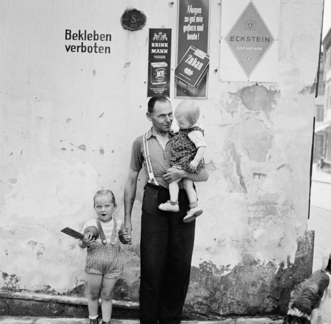 Отец с двумя детьми возле стенки на которой вопреки запрету висят рекламные плакаты Германия 1955 год